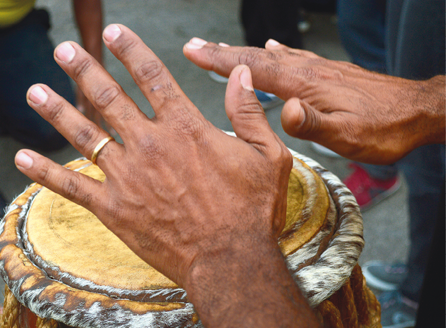 Fotografia. Vista do alto de mãos de uma pessoa morena batendo sobre bongô. Na mão esquerda, aliança dourada no dedo anelar. O instrumento, visto parcialmente, é similar a um tambor em tons de amarelo.
