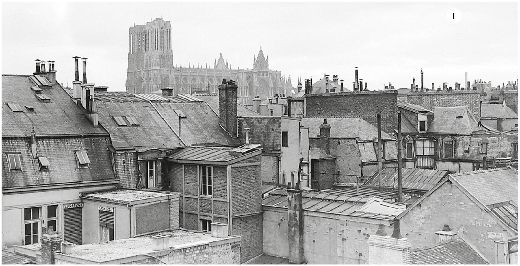 Fotografia um. Em preto e branco . Vista do alto de cidade francesa no final dos anos 30, com prédios pequenos entre residências. Ao fundo, uma catedral com duas torres à direita.