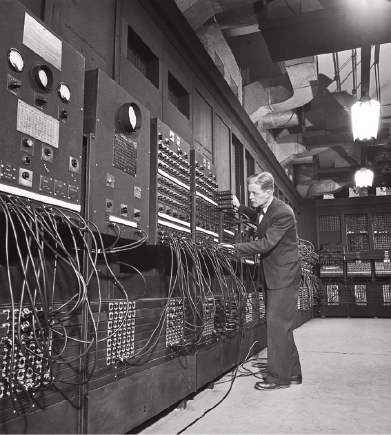 Fotografia. Em preto e branco. À esquerda, um computador grande escuro, com vários botões e fios conectados. À direita, um homem em pé, de frente para o computador, de terno e sapatos escuros.