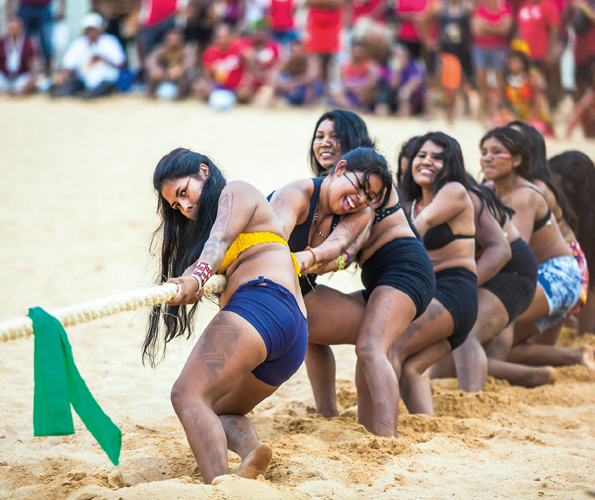 Fotografia. Em local com chão de areia, um grupo de mulheres puxando uma corda, com o corpo inclinado para trás. Elas são morenas, de cabelos longos pretos, usando top e um short. Em segundo plano, vista parcial de pessoas que as observam.