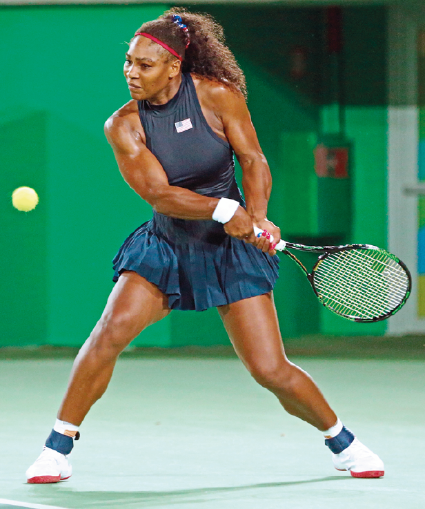 Fotografia. Em uma quadra de tênis, a jogadora de tênis Serena Williams, uma mulher negra, alta e musculosa, com cabelos longos em castanho, penteados e presos para trás, de regata e saia curta azul. Ela usa par de tênis branco e meia com detalhes em azul, segurando com as duas mãos uma raquete, para trás. Perto dela, uma bolinha de tênis amarela. Ao fundo, parede verde.