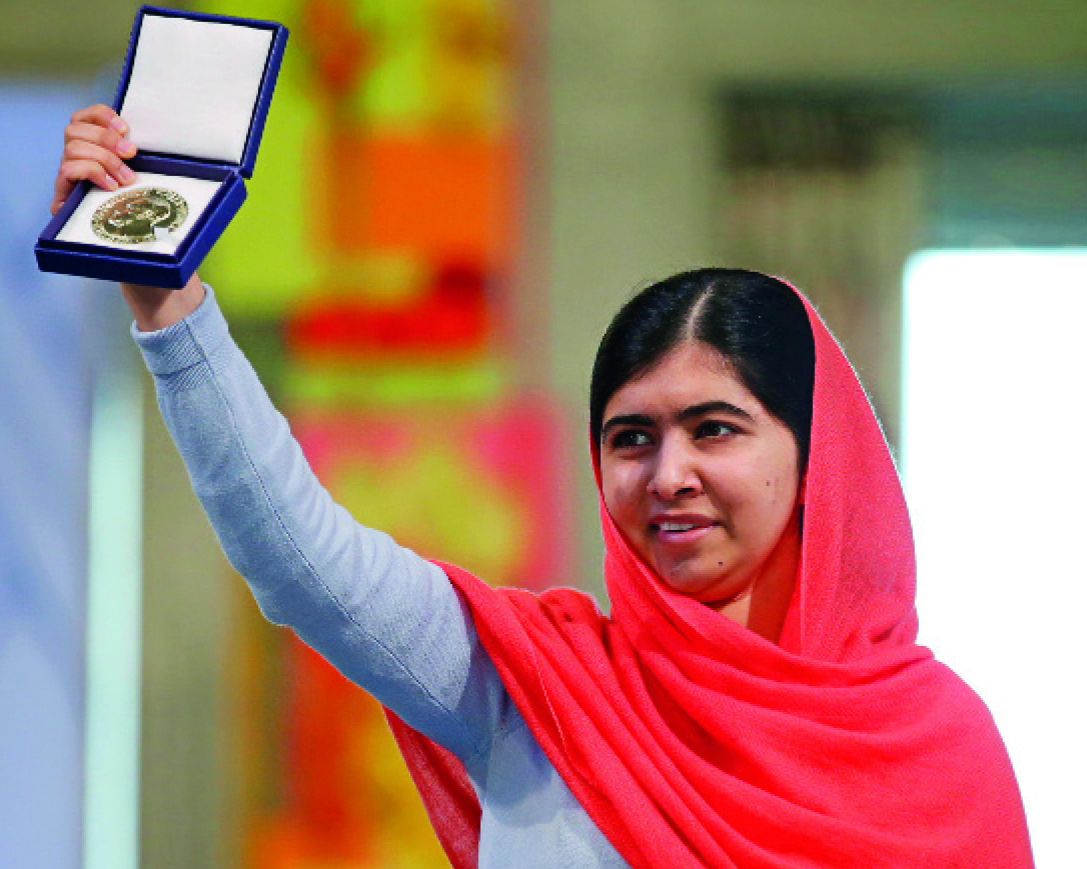 Fotografia. A ativista Malala Yousafzai, uma mulher vista dos ombros para cima, de cabelos pretos lisos penteados para a esquerda, sobrancelhas grossas e sobre a cabeça dela, um lenço vermelho que tampa os ombros. Ela está com o braço direito erguido para cima, mostrando uma caixa aberta azul com um objeto dentro dourado redondo.">