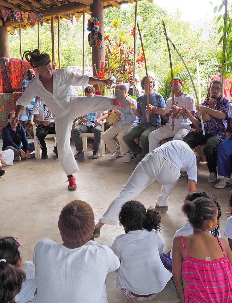 Fotografia. Em um local com piso claro, rosa de capoeira vista por pessoas e crianças sentadas ao redor. Algumas pessoas sentadas sobre o banco, à direita, segurando na mão um berimbau, instrumento com haste comprida em madeira envergada por um cabo de arame e com uma cabaça na parte de baixo.  Ao centro, duas pessoas dando golpes de capoeira, uma de frente para outra. À esquerda, homem moreno, de cabelos castanhos trançados para trás, com blusa de mangas três quartos e calça branca. Ele está equilibrado com a perna esquerda e perna direita esticada em um chute frontal. De frente para ele, outra pessoa vista de costas, com camiseta e calça brancas, corpo inclinado para a direita com as mãos encostando no chão e de cabelos escuros. Em segundo plano, local com vegetação e árvores de folhas verdes.