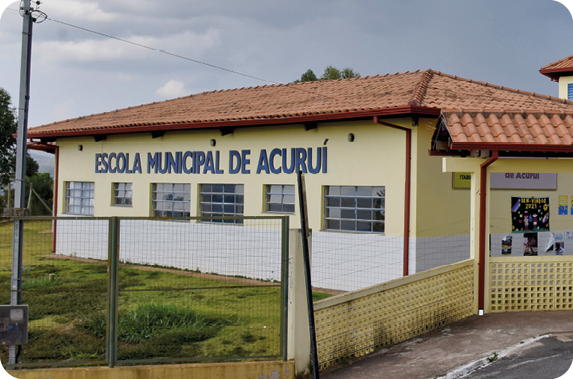 Fotografia. Vista geral de estabelecimento retangular, de paredes amarelas claro, telhado marrom, janelas e na parte superior, texto em azul: Escola Municipal de Acuruí. Ao redor, local com grama verde. Na parte superior, céu com algumas nuvens.