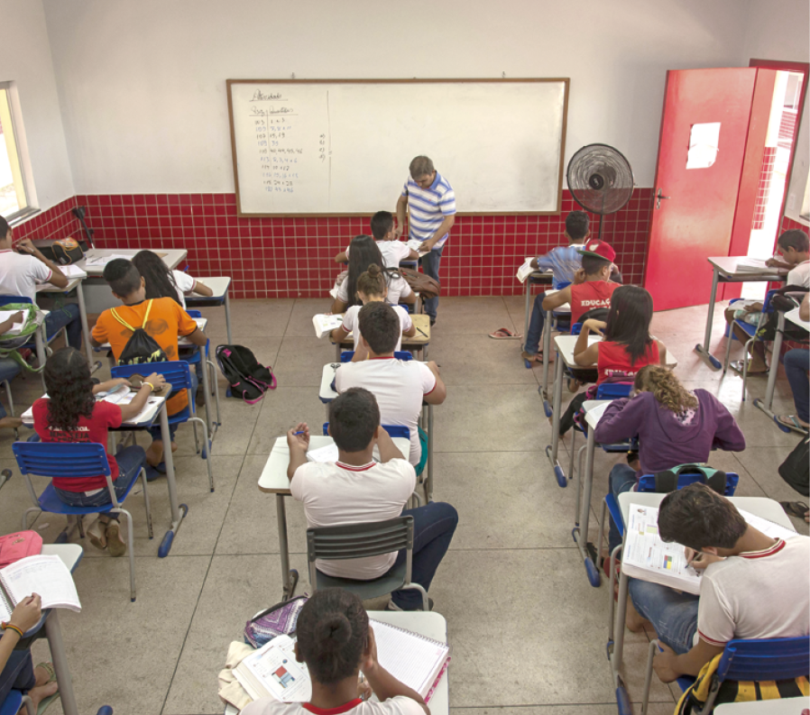 Fotografia. Vista geral de sala de aula vista de cima por trás das carteiras. Há três fileiras de alunos sentados em suas carteiras, olhando para a frente. Ao fundo, parede branca e vermelha, com lousa branca na horizontal. Perto de um aluno à frente, um homem em pé, de cabelos pretos e camiseta com faixas em branco e azul. Na ponta da direita, porta vermelha, aberta.