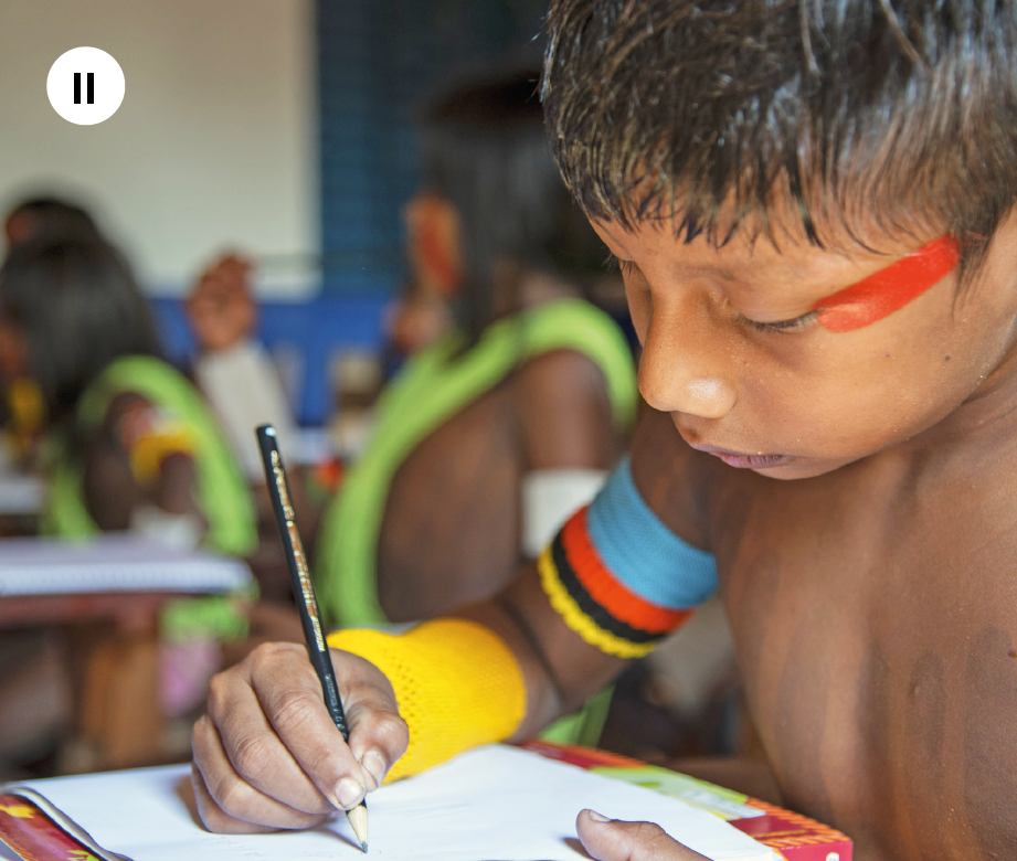 Fotografia dois. Menino indígena visto de lado, com pintura vermelha perto do olho esquerdo e adereços coloridos no braço direito. Ele escreve em um caderno. Ao fundo, há outras crianças indígenas em uma sala de aula.