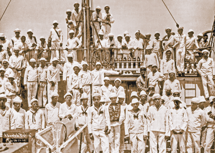 Fotografia. Em sépia, dezenas de homens em pé no convés de um navio, de roupas brancas de mangas compridas, calça e quepe, com lenço escuro ao redor do pescoço e pontas caídas no peito. Na parte superior, degrau com os outros homens em pé, perto do mastro ao centro.