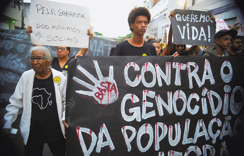 Fotografia. Em uma rua, pessoas se manifestando, em pé, algumas carregando cartazes. À frente, ao centro, menino de cabelos pretos curtos e camiseta preta. Na frente dele, cartaz preto com texto em branco: CONTRA O GENOCÍDIO DA POPULAÇÃO NEGRA. À esquerda, senhora de cabelos brancos penteados para trás, de blusa e calça pretas, com casaco branco de mangas comprida e par de óculos de grau. Ao fundo, à direita, pessoa vista parcialmente com um cartaz preto e texto em branco: QUEREMOS VIDA!