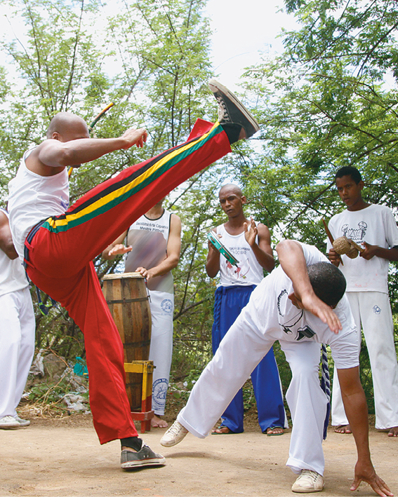 Fotografia. Dois homens um de frente para o outro, fazendo passos de capoeira. À esquerda, homem  com regata branca e calça vermelha, com listras finas na vertical em amarelo e verde, e tênis pretos. Ele está com o corpo equilibrado com a perna esquerda e a perna direita, esticada para a frente. À direita, um homem de cabelos pretos, com camiseta, calça e sapatos brancos, o corpo curvado para o seu lado esquerdo baixo, com perna esquerda no chão e a outra para trás, com braço esquerdo tocando o chão e o outro sobre o rosto. Em segundo plano, outras pessoas em pé e mais atrás, árvores de folhas verdes e céu claro.