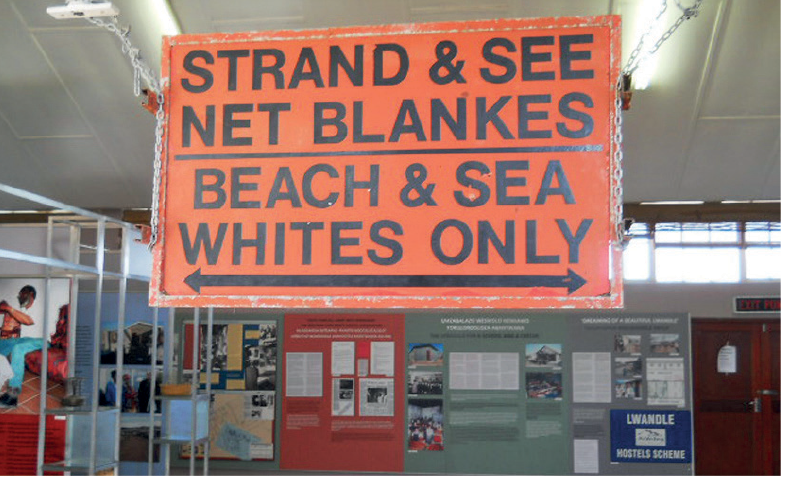 Fotografia. Vista geral de local fechado com paredes cinza e vários cartazes. Ao centro, na parte superior, faixa em laranja, com texto em preto: STRAND & SEE NET BLANKES BEACH & SEA WHITES ONLY.