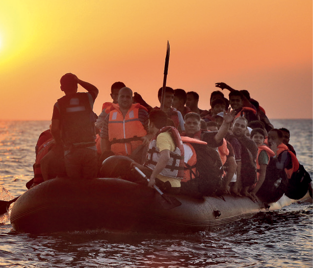Fotografia. Em alto-mar, um bote inflável com dezenas de pessoas sobre ele. Muitas estão em pé, algumas com colete salva-vidas em laranja. Em segundo plano, céu em tons de laranja e sol à esquerda.