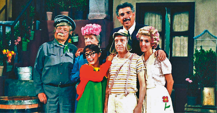 Fotografia. Turma da série de TV Chaves. Seis pessoas distribuídas em duas fileiras de três, uma na frente da outra. Eles estão em frente a uma vila com janelas marrons e, à esquerda, escada com corrimão e balde cinza sobre um barril. Da esquerda para direita, um senhor com bigode branco, de boina, blusa de mangas compridas e calça cinza; uma mulher de cabelos grisalhos, com flores rosa sobre a cabeça e blusa azul de mangas compridas, parcialmente encoberta pela menina que está à sua frente. Mais à direita, um homem alto de cabelos e bigode grisalhos, de camisa branca e terno cinza. À frente deles, uma garota de cabelos escuros, colete vermelho sobre vestido verde, par de óculos de grau preto. Ele tem o braço esquerdo sobre ombro de um garoto com gorro com abas laterais que cobrem as orelhas, camiseta de listras em bege e marrom, com jardineira em bege com duas alças finas sobre o ombro direito. E mais à direita mulher de cabelos enrolados com bobes de cabelo, de blusa em bege e avental claro na cintura. Ambos olham para frente, posando para foto.