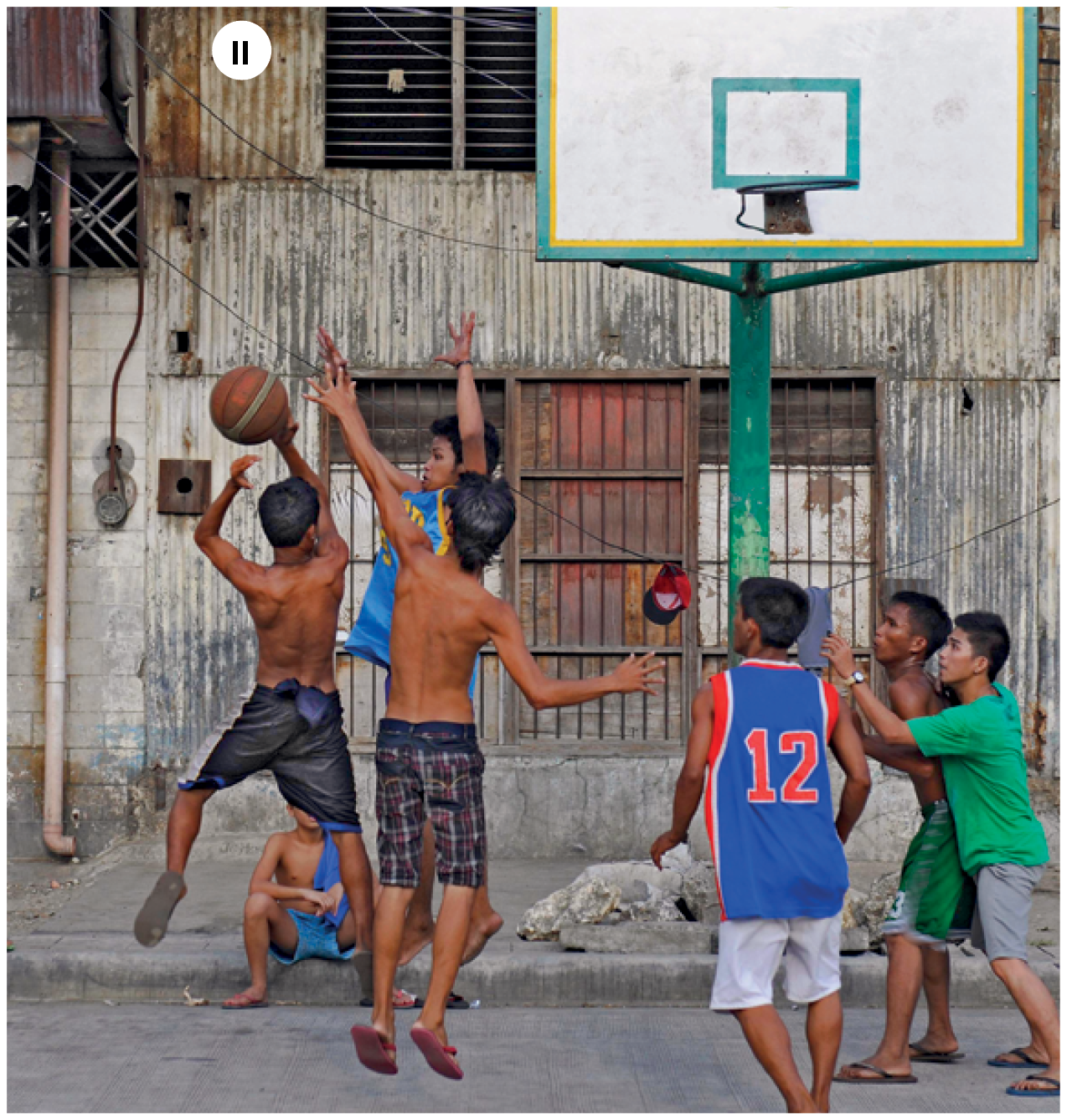 Fotografia dois. Vista geral de uma rua asfaltada, com seis garotos jogando basquete, sendo que três deles estão sem camisetas, perto de uma tabela e cesta com haste verde, com parte superior em branco e contornos em verde e amarelo, que é mantida em pé com a ajuda de grandes pedras na base. Três jogadores estão à esquerda disputando uma bola que está no alto. À direita, os outros três jogadores observam. Em segundo plano, prédios de parede de madeira.