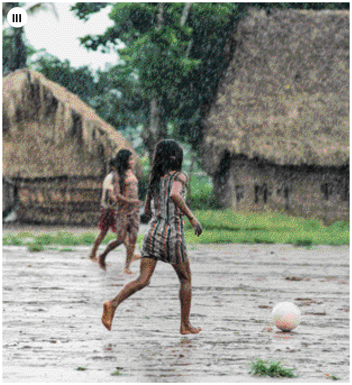 Fotografia três. Três meninas brincando com bola branca, com local aberto. Chão de terra com trechos com grama verde e casas de palhas em bege entre árvores de folhas verdes ao fundo. À frente, menina de cabelos escuros, com vestido curto com listras verticais, correndo para a direita atrás de uma bola. Ao fundo, outras duas meninas.