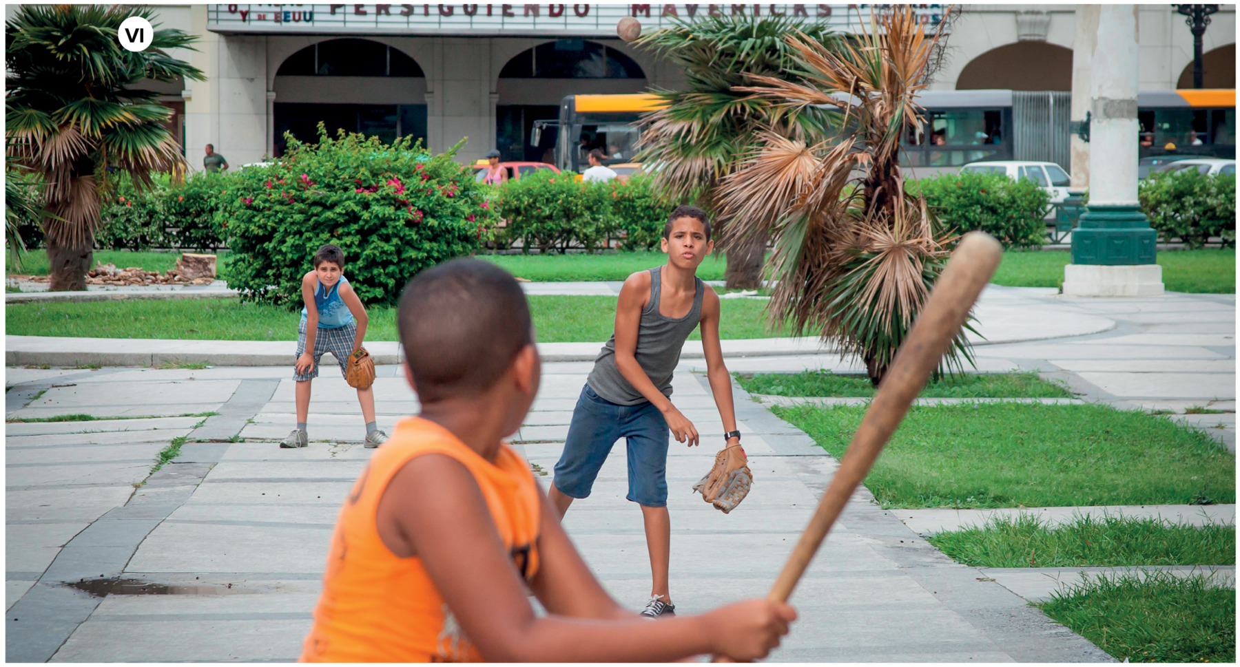 Fotografia seis. Em um local aberto, aparentemente uma praça em uma cidade, há três meninos jogando beisebol. Um dos meninos está de costas, da cintura para cima, de cabelos pretos curtos, com regata em laranja. Este segura nas mãos, um bastão de beisebol feito de madeira. Mais à frente, um menino em pé, olhando para frente, com uma luva de beisebol de couro na mão esquerda, com regata cinza, calça jeans azul e sapatos cinza. Ele tem cabelos castanhos. Mais ao fundo, à esquerda, um outro menino com o corpo um pouco curvado para frente, com as mãos sobre os joelhos. Ele tem cabelos castanhos, usa blusa regata azul, bermuda xadrez cinza e sapatos bege. Em segundo plano, local com grama verde, árvores, arbustos e plantas ornamentais.  Mais ao fundo, um prédio com várias portas grandes em arco.