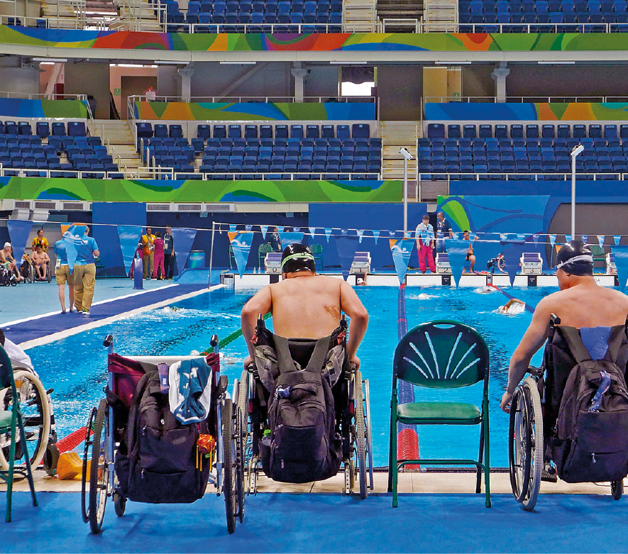 Fotografia. Vista geral de um ginásio de natação com uma piscina de água azul. De frente para ela, cadeiras e dois  homens vistos de costas, sentados em suas cadeiras de rodas com mochila penduradas na parte de atrás. Ambos têm  cabelos escuros e usam touca de natação e óculos. Ao fundo, as arquibancadas do ginásio em azul.