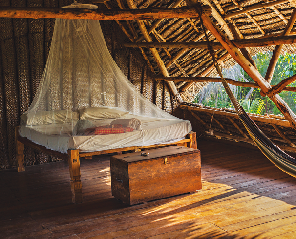 Fotografia. Interior de uma cabana de madeira com uma cama coberta por mosquiteiro de cor branca. À frente da cama, uma mobília de madeira marrom, similar a um pequeno baú. À direita, abertura por onde vê-se folhas verdes de árvores.