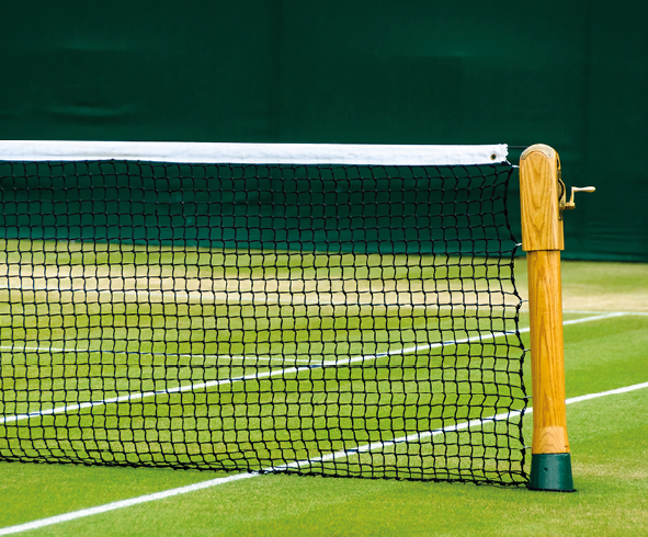 Fotografia. Vista parcial de quadra de tênis com grama verde, linhas em branco e uma rede na horizontal esticada com hastes amarelas na vertical à direita.