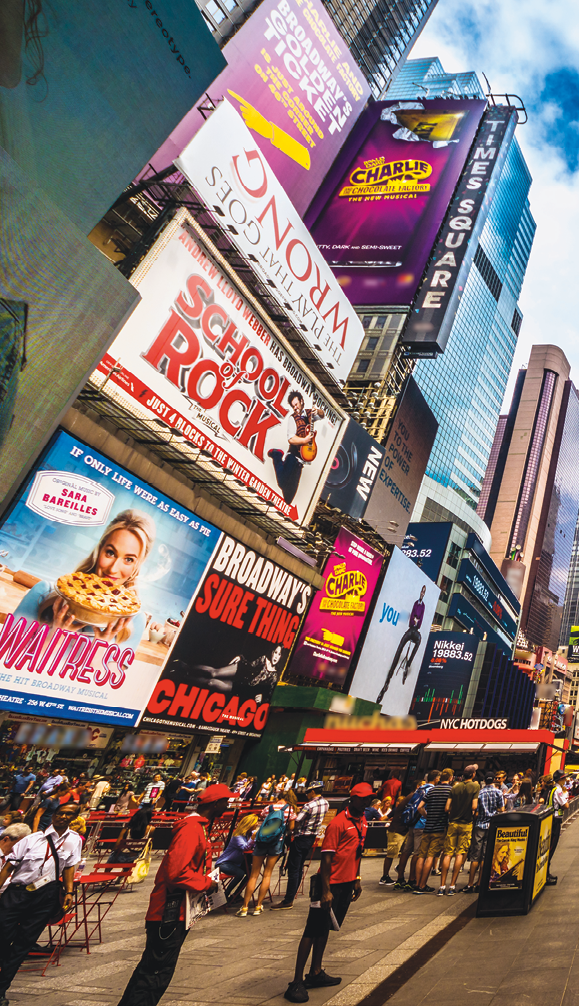 Fotografia. Exterior de uma rua movimentada, onde há várias pessoas andando na calçada e ao fundo, do lado esquerdo, há fachadas de prédios altos e cartazes anunciando diversos shows de teatro e musicais como Chicago, School of Rock, Waitress e outros.