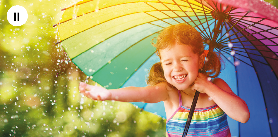 Fotografia. Uma criança em área externa onde chove. Ela segura um guarda-chuvas multicolorido nas cores azul, verde, amarelo, roxo e outras. Ela tem os cabeços presos nas laterais da cabeça com elásticos. Ela veste um vestido de alças finas com listras horizontais em várias cores. Ela tem o braço direito esticado sentindo a chuva com a palma das mãos.