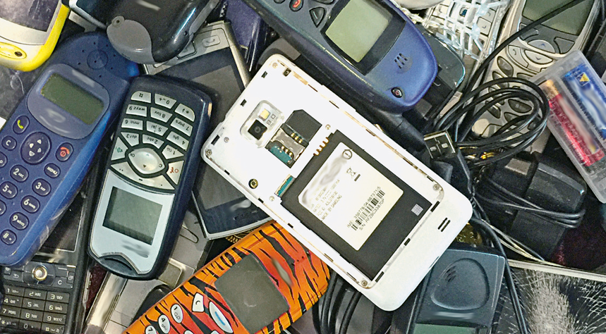 Fotografia. Vista do alto de vários aparelhos eletrônicos descartados, um sobre o outro, de telefones, celulares e entre outros, sobre fios pretos.