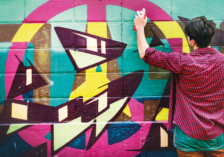 Fotografia. Uma parede com graffiti abstrato em rosa, amarelo, roxo e branco. À direita, um homem visto de costas, de cabelos pretos, de camisa de mangas arregaçadas em rosa e roupa de baixo em verde. Ele segura na mão esquerda um tubo de spray de frente para parede.