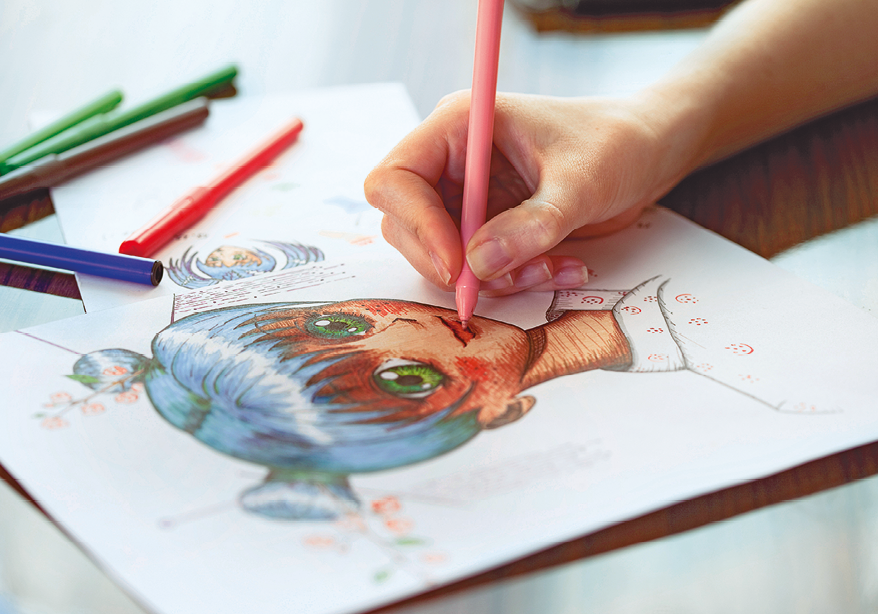 Fotografia. Mão de uma mulher, segurando caneta colorida rosa. Ela colore a boca de uma menina que está desenhada na folha de papel branco. À esquerda, outras canetas coloridas.