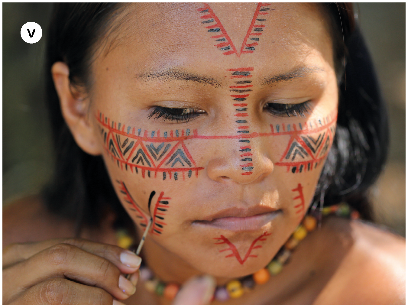 Fotografia. Número cinco. Menina indígena fazendo pintura em seu rosto com a ajuda de um graveto fino na mão direita. Os desenhos são nas cores vermelha e preta e têm motivos geométricos.