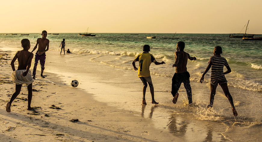 Fotografia. Vista geral de uma praia com mar à direita. Na areia, cinco meninos jogando futebol, com uma bola à esquerda.