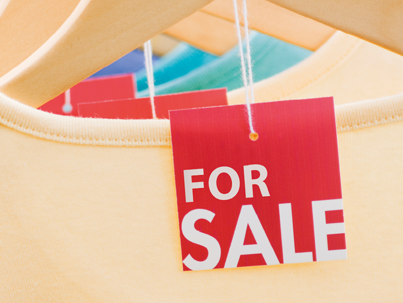 Ilustração: close de camisetas amarelas, azuis e verdes penduradas em cabides com destaque para as etiquetas vermelhas com o texto: for sale.