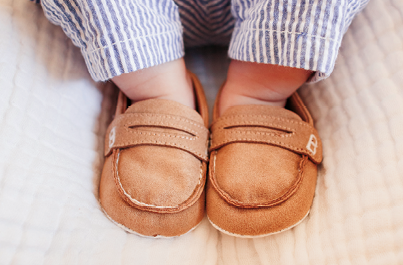 Fotografia. Foco nos pés de um bebê calçando mocassins marrons velhos e laceados com calça listrada na vertical em branco e azul.
