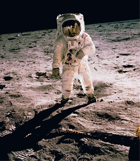 Fotografia. Um astronauta, de roupa branca, com capacete com visor espelhado, par de luvas cinzas, caminhando sobre um solo arenoso em bege. Ao fundo, céu de cor preta.