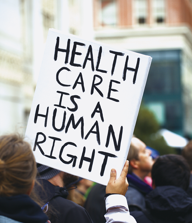Fotografia. Uma pessoa vista de costas, dos ombros para cima, de blusa em preto e branco. Na mão direita, segura um cartaz branco com texto em preto: Health care is a human right. Ao fundo, vista parcial de pessoas e prédios.
