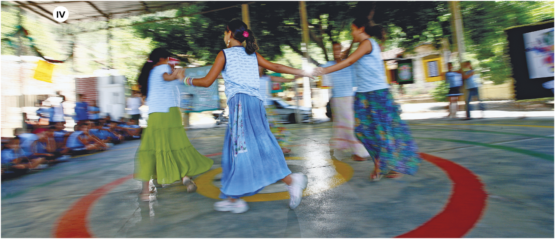 Fotografia. Número quatro. Quatro mulheres dançando em pé, de mãos dadas, fazendo ciranda, em local com solo em azul e círculos coloridos em verde, vermelho e amarelo. As mulheres têm cabelos escuros presos para trás, de blusa regata em azul-claro e saiotes longos. Em segundo plano, à esquerda, crianças sentadas. Ao fundo, árvores e outras pessoas desfocadas.