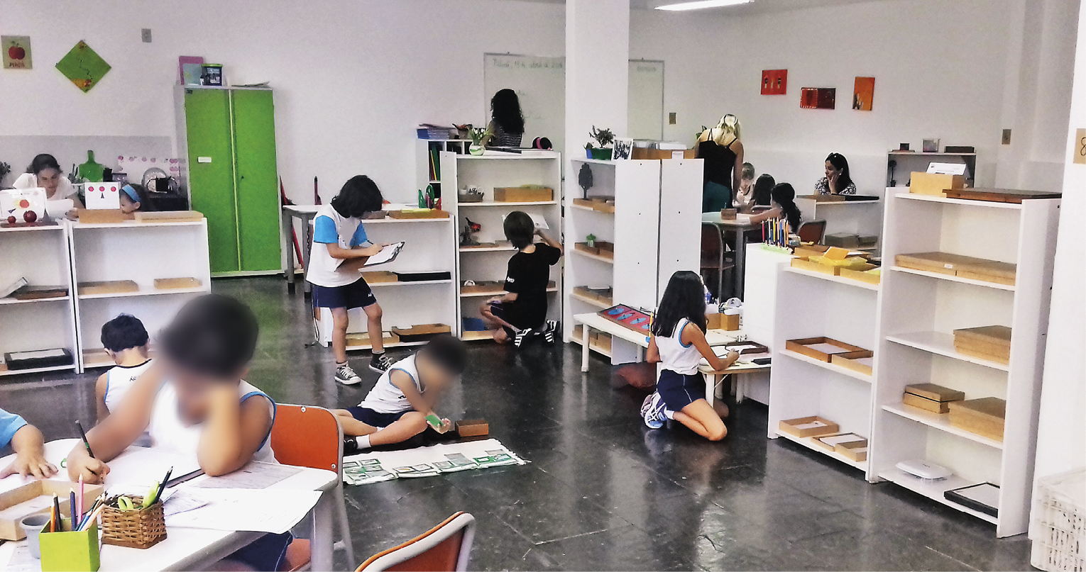 Fotografia. Sala de aula com paredes em branco e piso em cinza. Ao redor, estantes pequenas em branco, com caixas de madeira parecidas com estojos, com crianças agachadas perto e à esquerda, outras crianças de frente para mesa. Em segundo plano, um armário de portas em verde.