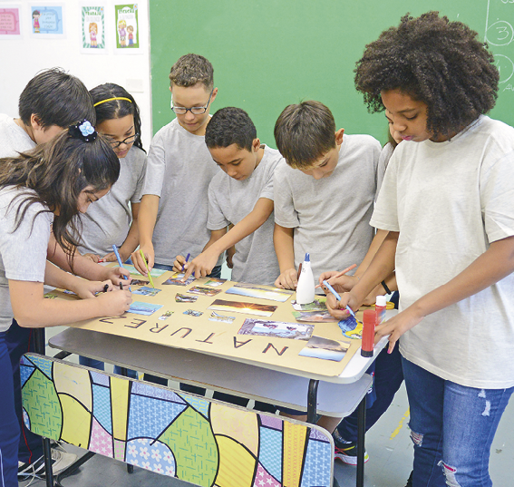 Fotografia. Vista geral de uma mesa na horizontal fazendo um cartaz juntas. Meninos e meninas de camiseta cinza e calça azul. Elas estão com a cabeça para baixo, com utensílios nas mãos, criando o cartaz.