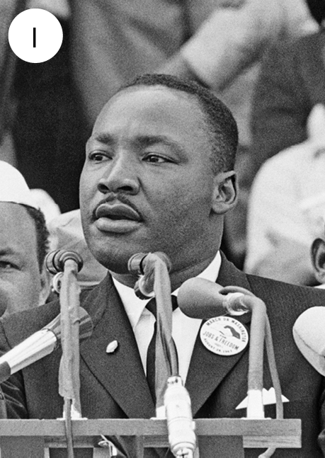 Fotografia. Número um.  Em preto e branco. O ativista Martin Luther King Jr, visto do busto para cima, de frente para microfones sobre uma base. Ele tem  cabelos e bigode escuros, de camisa de gola clara, gravata e terno escuro, olhando para a esquerda, com a boca um pouco aberta.