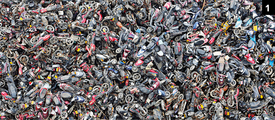 Fotografia. Número um. Vista do alto de um ferro-velho com destroços de motocicletas empilhadas, de cor cinza e outras em tons de vermelha.