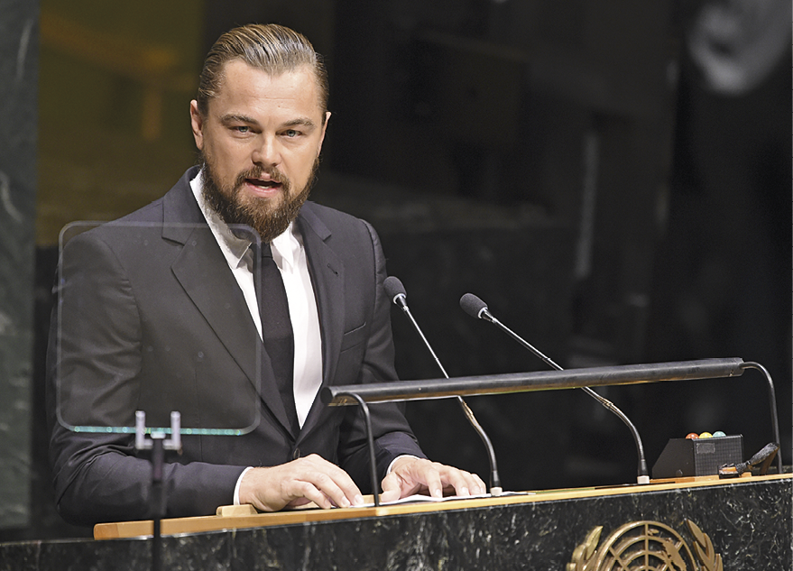 Fotografia. O ator Leonardo DiCaprio, de frente para púlpito preto, visto da cintura para cima. Ele tem cabelos e barba claros, usa camisa de gola branca, com gravata e terno em cinza. O ator fala perto de dois microfones pequenos sobre um púlpito com o símbolo das Nações Unidas em dourado.