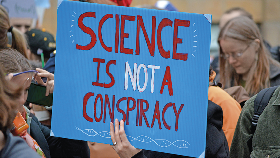 Fotografia. Uma pessoa segurando um cartaz em azul-claro e texto: Science is not a conspiracy. Ao fundo, pessoas em pé, aglomeradas, vistas parcialmente.