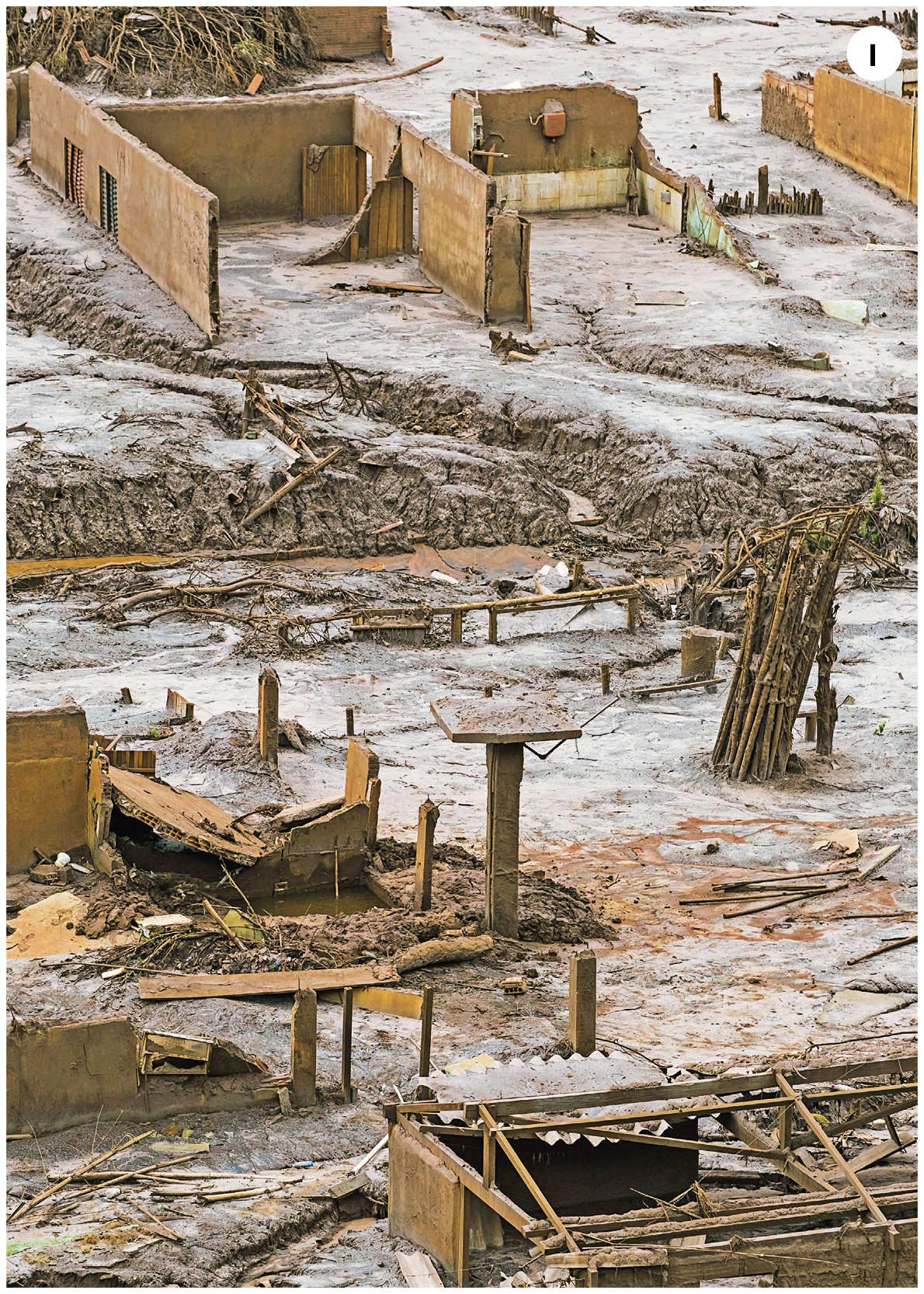 Fotografia. Número um. Vista do alto de local com casas destruídas, com restos de paredes e vigas de madeira, de cor marrom. Sobre o solo e os destroços, neve branca.