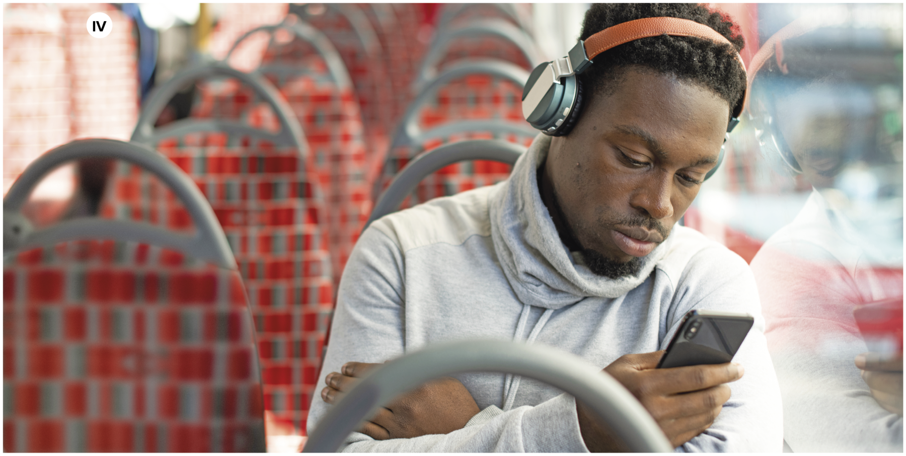 Fotografia. Número quatro.  Um homem visto da cintura para cima, dentro de um ônibus com assentos quadriculados em cinza e vermelho, olhando para um celular preto na mão direita. Ele usa um fone sem fio cobrindo os ouvidos totalmente em vermelho e preto e usa blusa de frio de mangas compridas e gola alta em azul claro, com os braços cruzados.