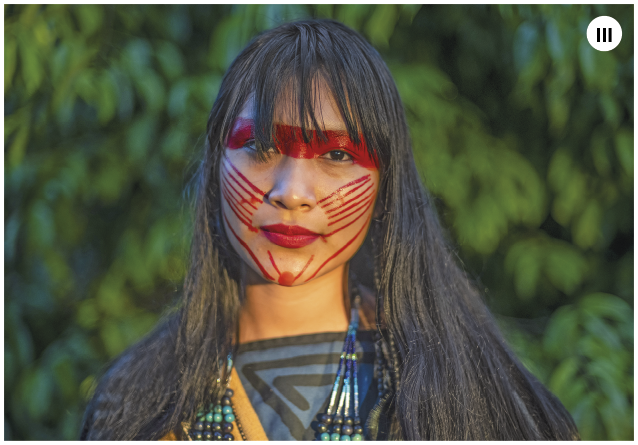 Fotografia. Número três.  Uma mulher indígena vista dos ombros para cima. Ela tem cabelos longos pretos com franja. Tem uma pintura em forma de  faixa horizontal em vermelho em cima dos olhos, e linhas finas sobre as bochechas. Usa um tecido sobre os ombros em azul e preto e um colar. Ao fundo, vegetação em verde.