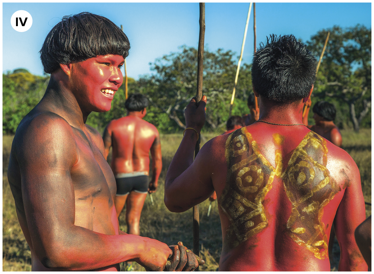 Fotografia. Número quatro. Grupo de homens indígenas vistos da cintura para cima, de cabelos escuros e lisos, com pinturas pelo corpo em tons de vermelho, preto e amarelo. Alguns levam lanças nas mãos. No primeiro plano, um dos homens está sorrindo. Ele tem uma pintura em vermelho no rosto. Os outros estão de costas. Ao fundo, vegetação verde. No alto, céu em azul-claro.