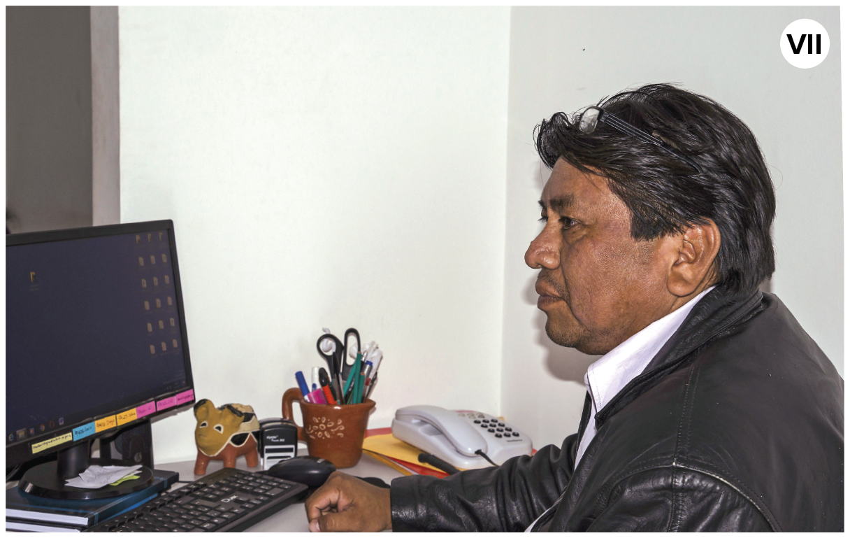 Fotografia. Número sete.  Um homem indígena está sentado de frente para uma mesa, onde há um computador preto. Ele tem cabelos pretos lisos e usa casaco de cor preta e camisa de gola em branco.