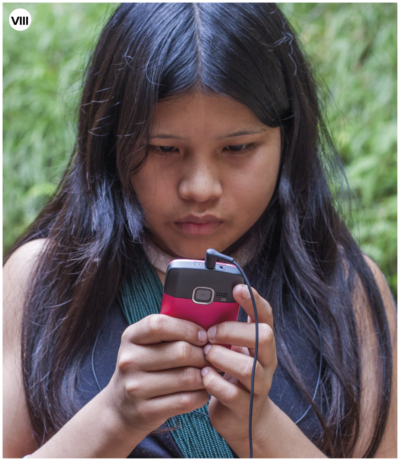 Fotografia. Número oito.  Uma menina indígena vista dos ombros para cima. Ela tem cabelos pretos longos e divididos ao meio. Ela segura nas mãos um celular em preto e rosa-escuro, e fones de ouvido. Ao fundo, vegetação com folhas em verde."