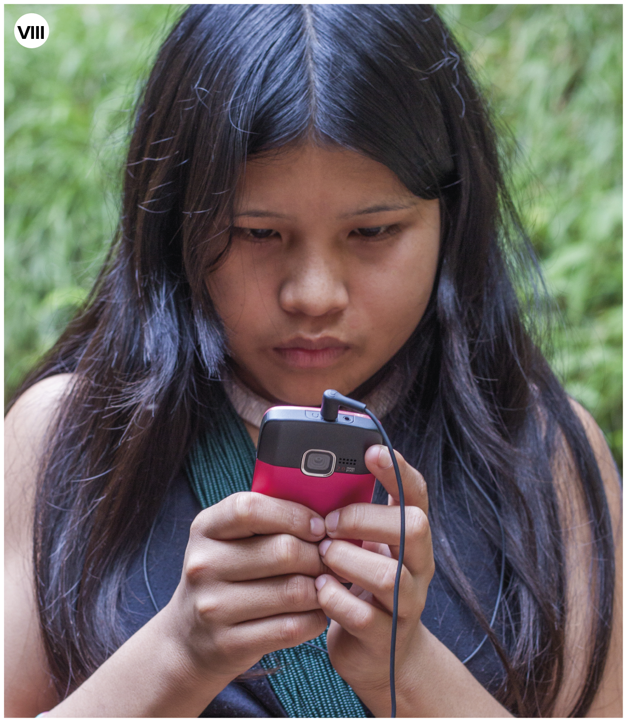 Fotografia. Número oito. Uma menina indígena vista dos ombros para cima. Ela tem cabelos pretos longos e divididos ao meio. Ela segura nas mãos um celular em preto e rosa-escuro, e fones de ouvido. Ao fundo, vegetação com folhas em verde."