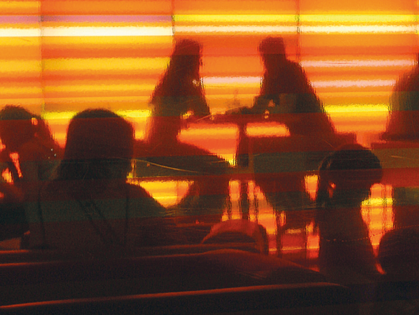Fotografia. Silhuetas de pessoas dentro de uma sala vistas através de uma persiana meio transparente, em tons de amarelo, laranja e vermelho. Há mulher sentada de frente para um homem. Ao redor, outras pessoas sentadas de frente para mesas.