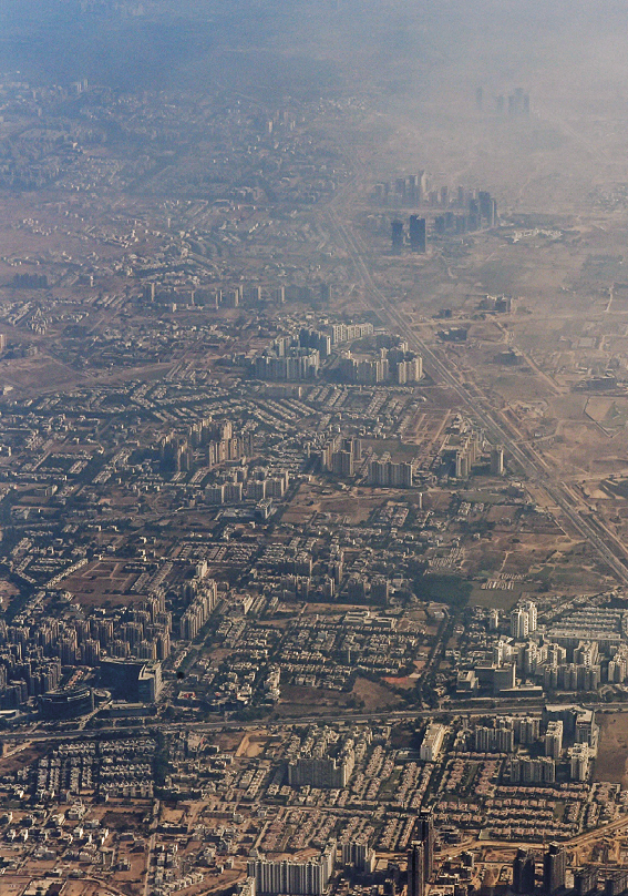 Fotografia. Vista aérea de uma grande cidade com muitas casas e alguns prédios. Uma fumaça acinzentada cobre grande parte da cidade.