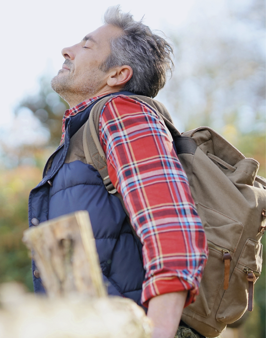 Fotografia. Um homem visto da cintura para cima, com o corpo para a esquerda. Ele tem cabelos grisalhos e barba rala, de olhos fechados, sorrindo, de camisa quadriculada vermelha e detalhes em azul com as mangas arregaçadas e colete acolchoado em azul. Sobre as costas, uma mochila em marrom. Perto dele, vista parcial de pedaços de madeira em bege-claro. Em segundo plano, vegetação seca desfocado.